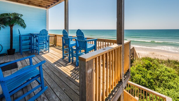 Terraza de madera con sillas Adirondack azules y una mesa, con vista a una playa.