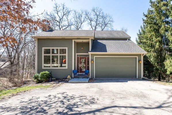 Une maison à deux étages avec un garage, un porche à l’avant doté de deux chaises et un petit jardin devant la maison.