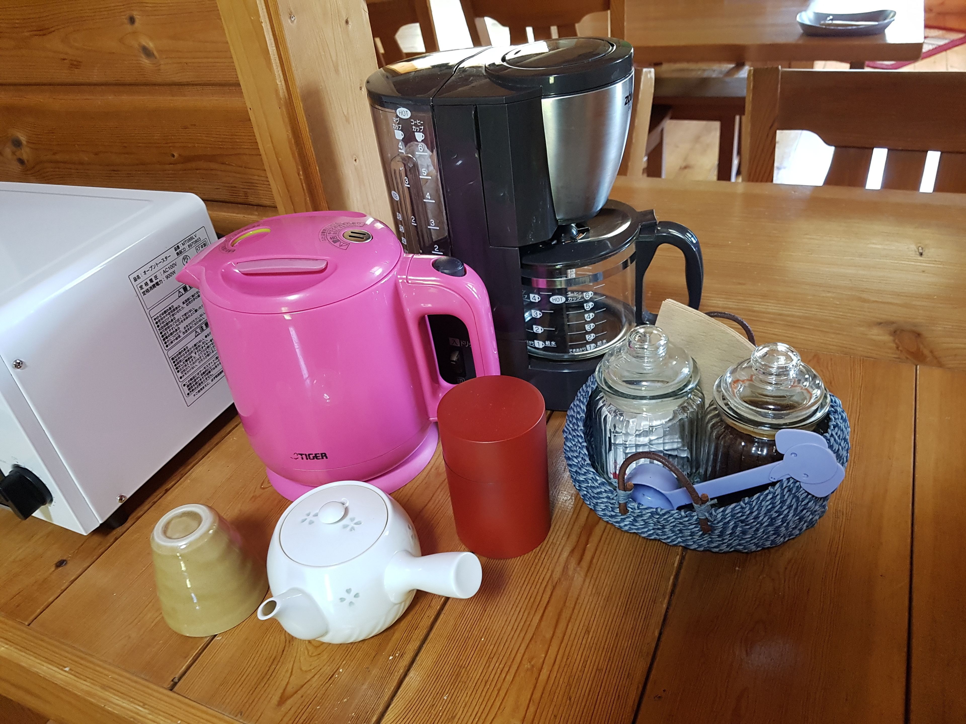 A kitchen with a pink electric kettle, a coffee maker, a white teapot, a red mug, and a glass jar with a lid.
