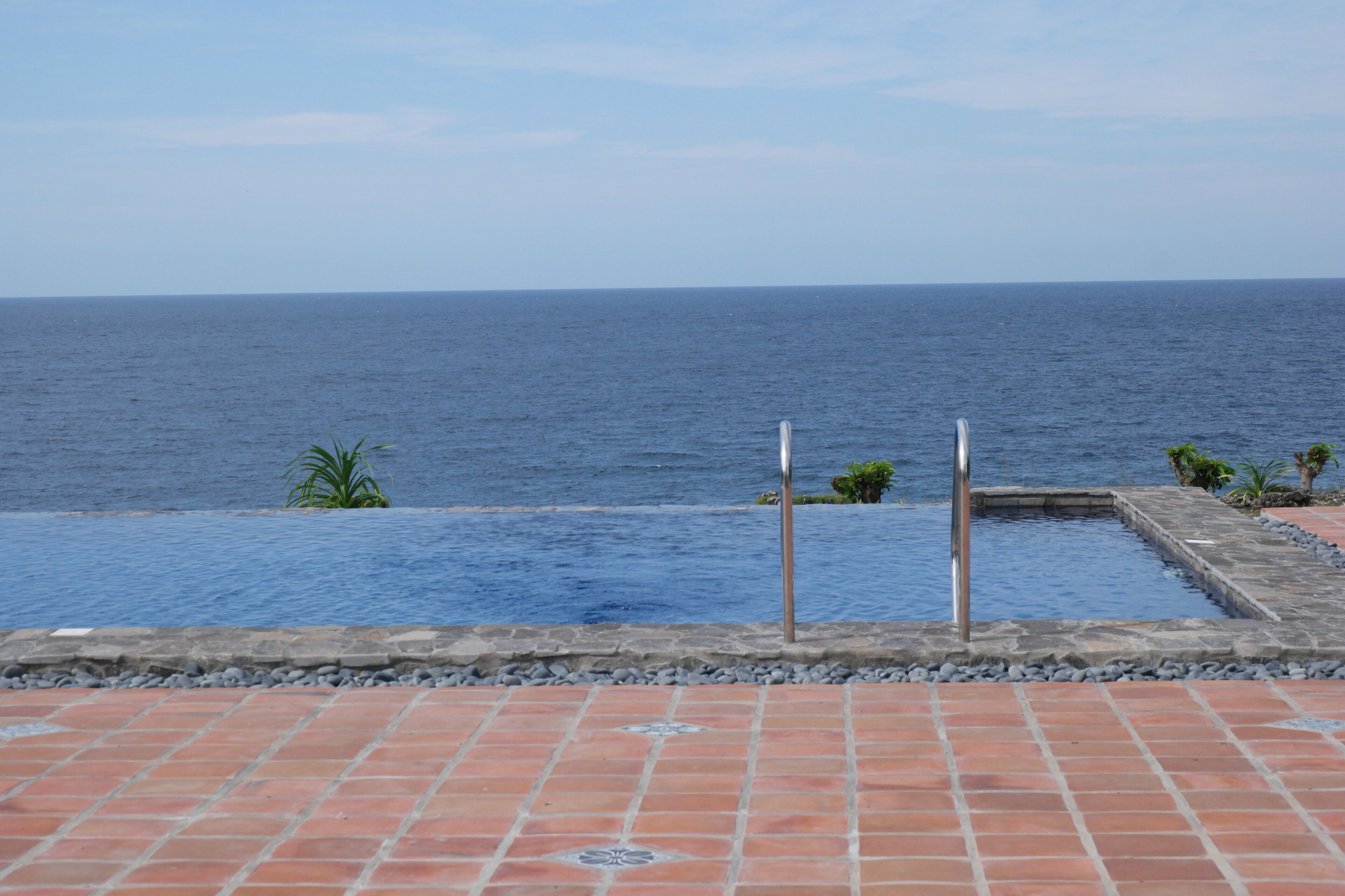 A pool with a view of the ocean, surrounded by a tiled patio.