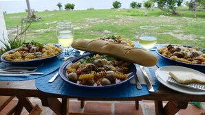 Una mesa dispuesta con platos de pasta, copas de vino blanco y una baguette, con vistas a una zona verde con árboles y el mar.