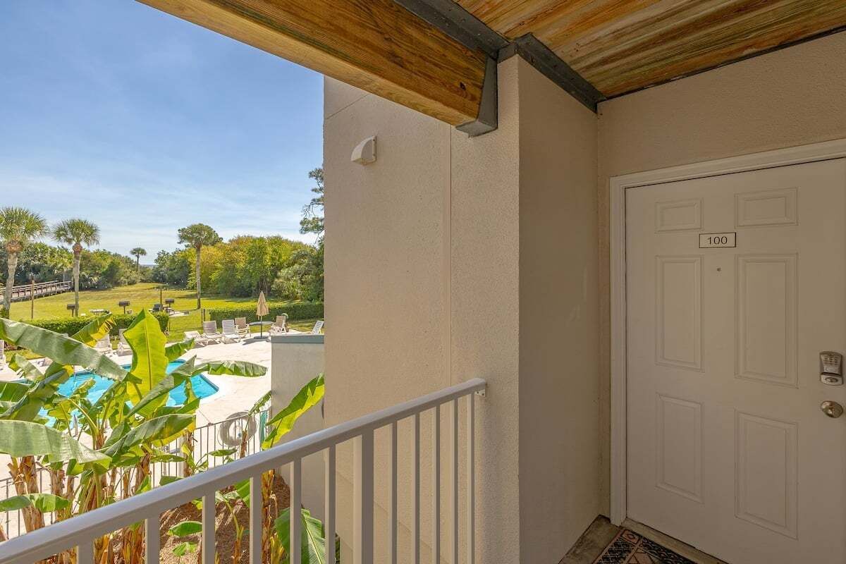 Un balcón con vista a la piscina, una puerta blanca con un número y una pequeña zona ajardinada.