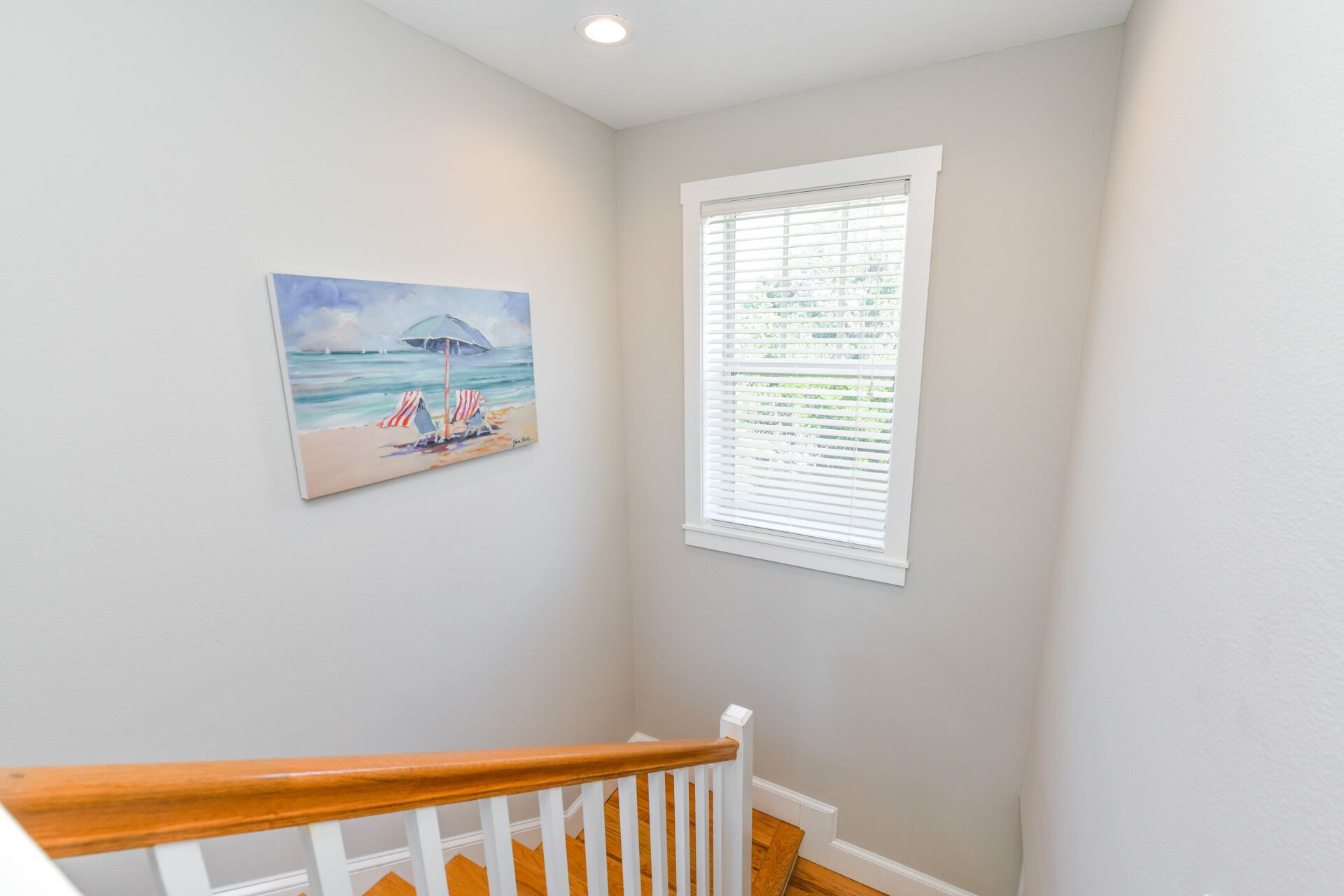 A staircase with a wooden handrail and white balusters, leading to a room with a beach-themed painting and a window with blinds.