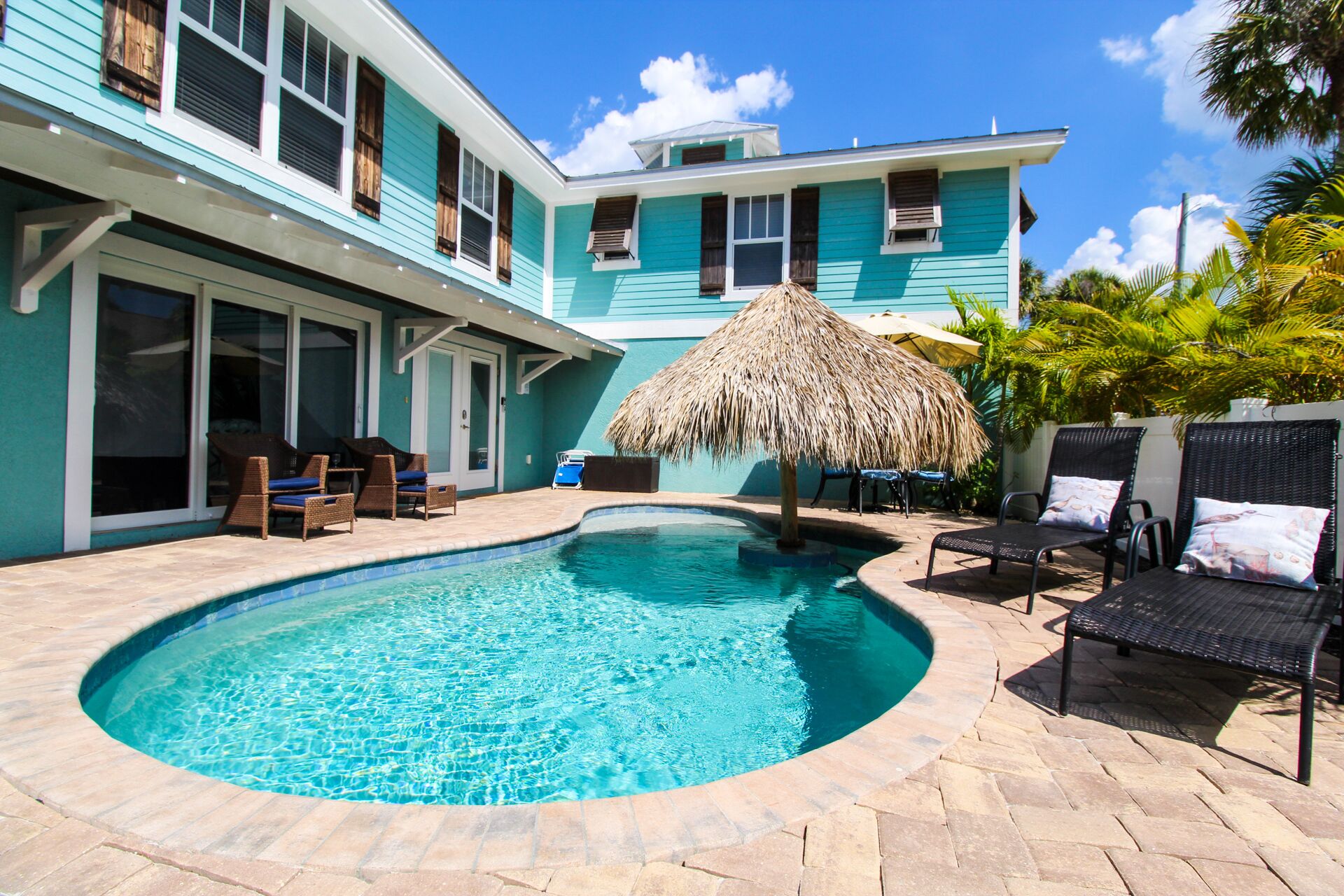 A pool area with lounge chairs and a thatched umbrella, surrounded by a two-story building with large windows.