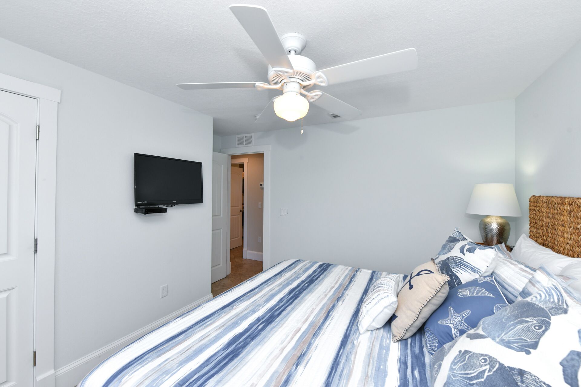 A bedroom with a bed, a ceiling fan, a TV mounted on the wall, and a lamp on the bedside table.