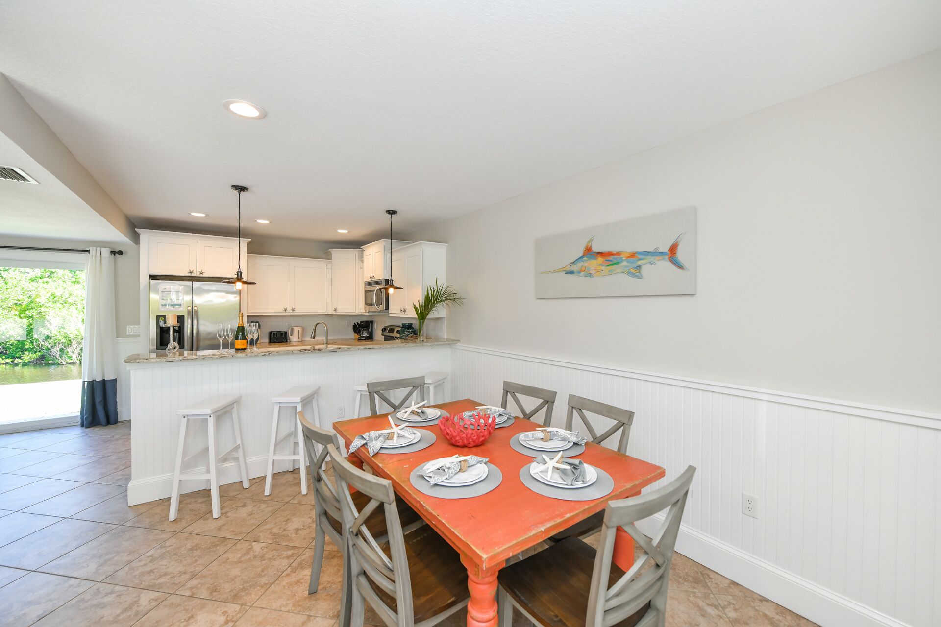 A dining area with a wooden table set for four, white bar stools, a kitchen in the background, and a colorful fish painting on the wall.