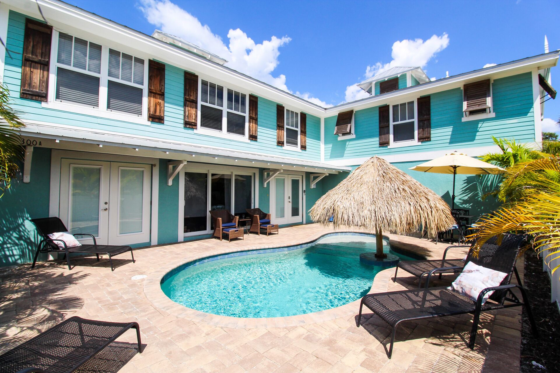 A two-story building with a swimming pool, outdoor seating, and a thatched umbrella.