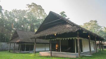 A thatched-roof structure with a porch and a covered seating area, surrounded by lush greenery.