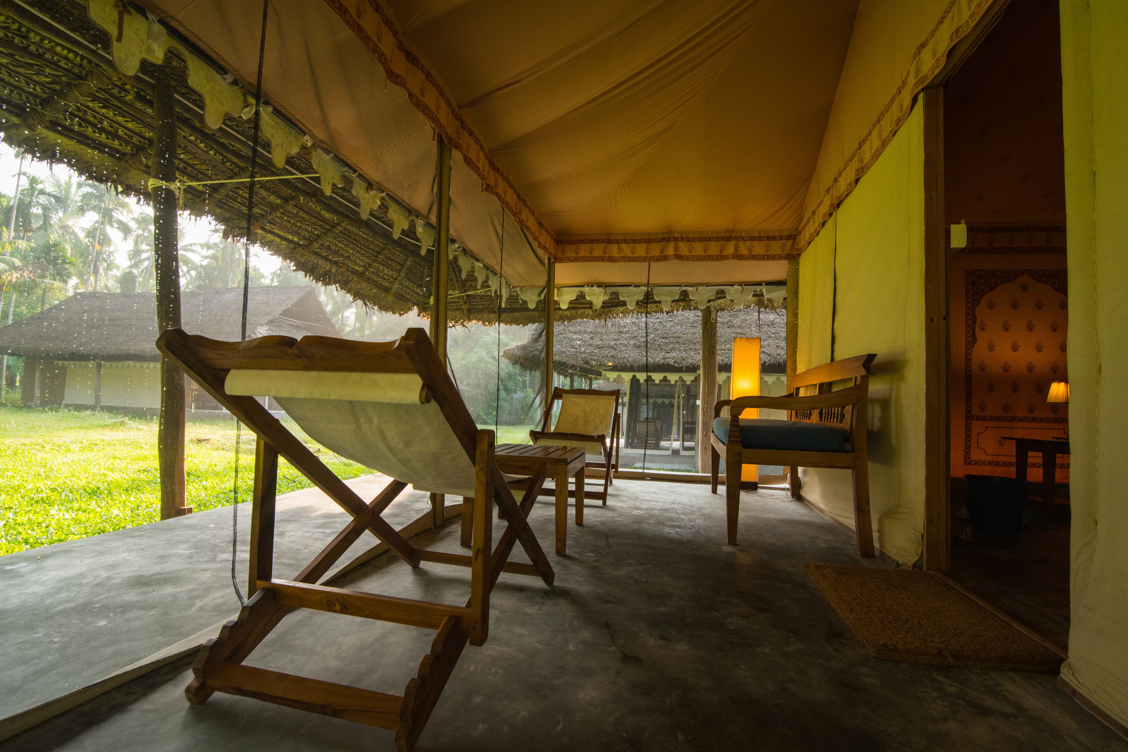 A covered outdoor seating area with wooden chairs and a bench, overlooking a garden with a thatched-roof structure.