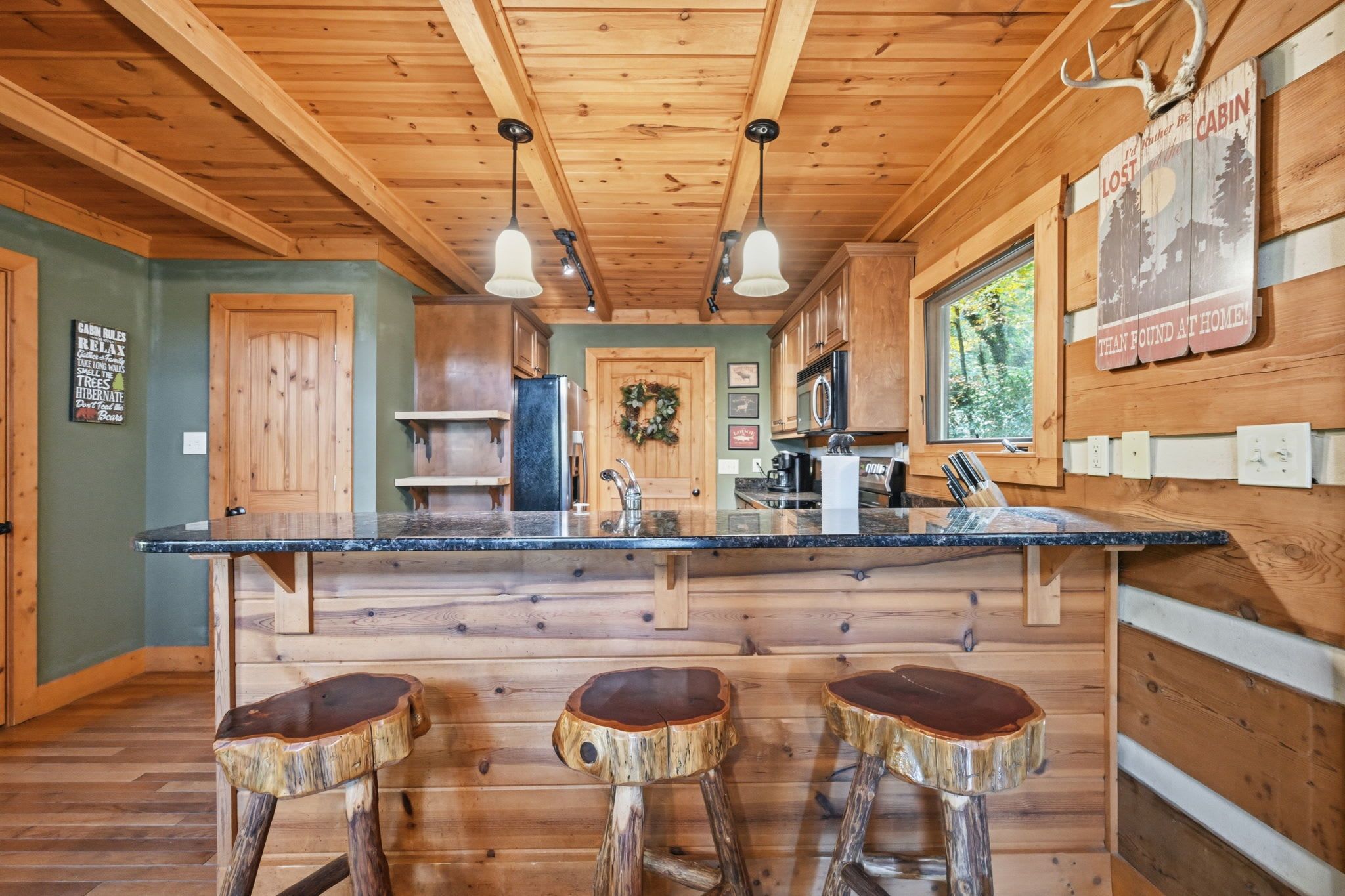 A cabin interior with a wooden bar area, a refrigerator, a microwave, and a window overlooking greenery.
