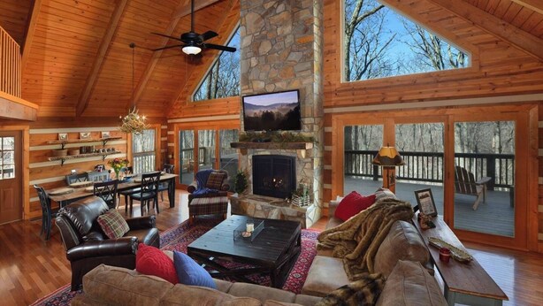 A spacious living room with a stone fireplace, wooden ceiling, and large windows overlooking a deck. - Luxury Cabin: 3 Kings, Hot Tub, Fireplace, Lg Deck (Banner Elk)