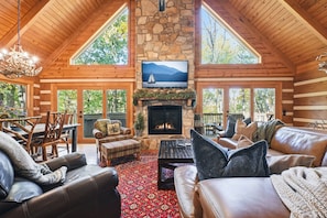 A log cabin interior with a stone fireplace, wooden furniture, and a large TV screen.
