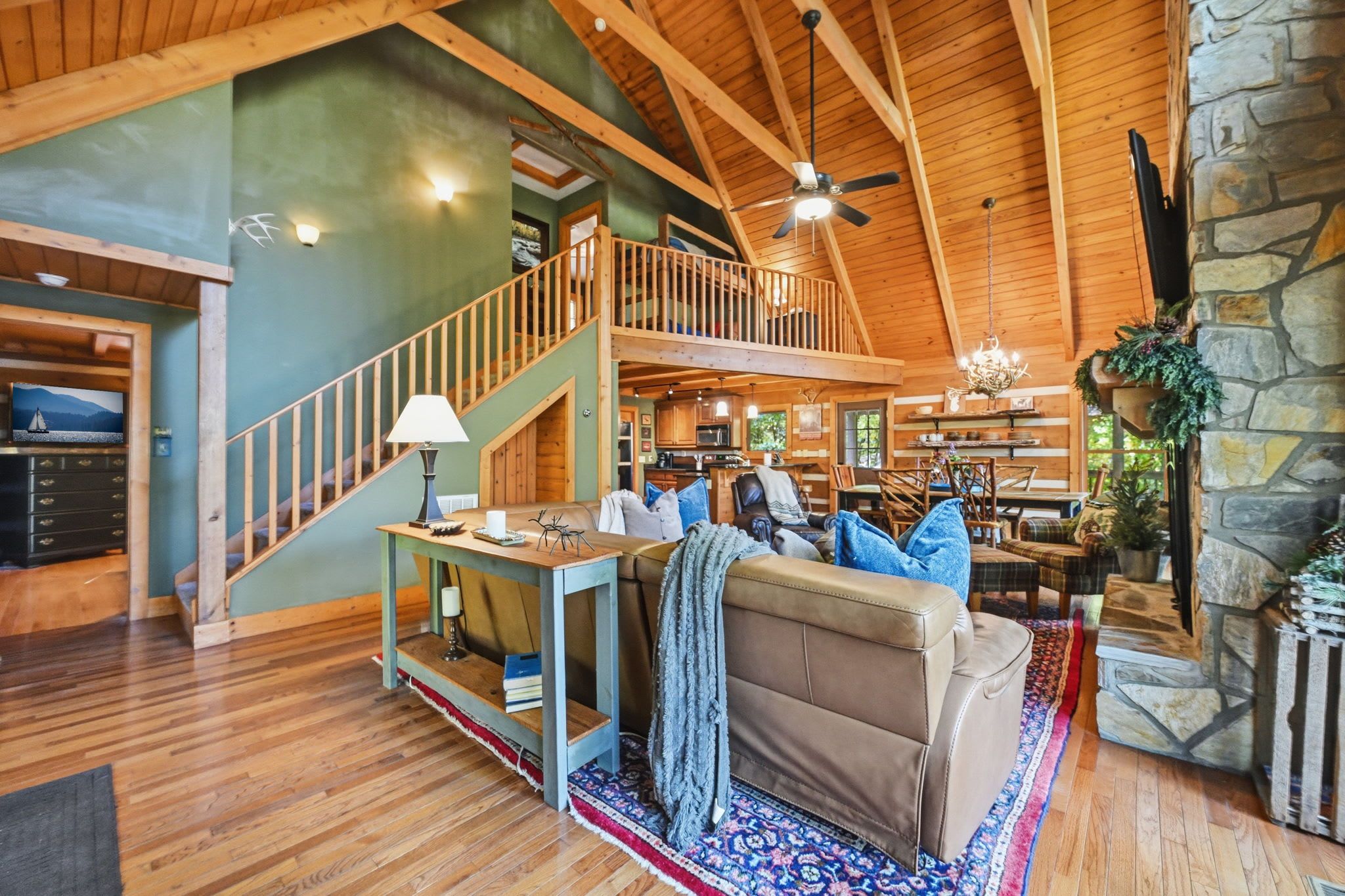 A spacious living area with a high ceiling, wooden beams, a staircase, a fireplace, and a cozy seating arrangement.