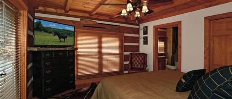 A bedroom with a wooden ceiling, a television on a dresser, a bed with a beige blanket, and a window with blinds.
