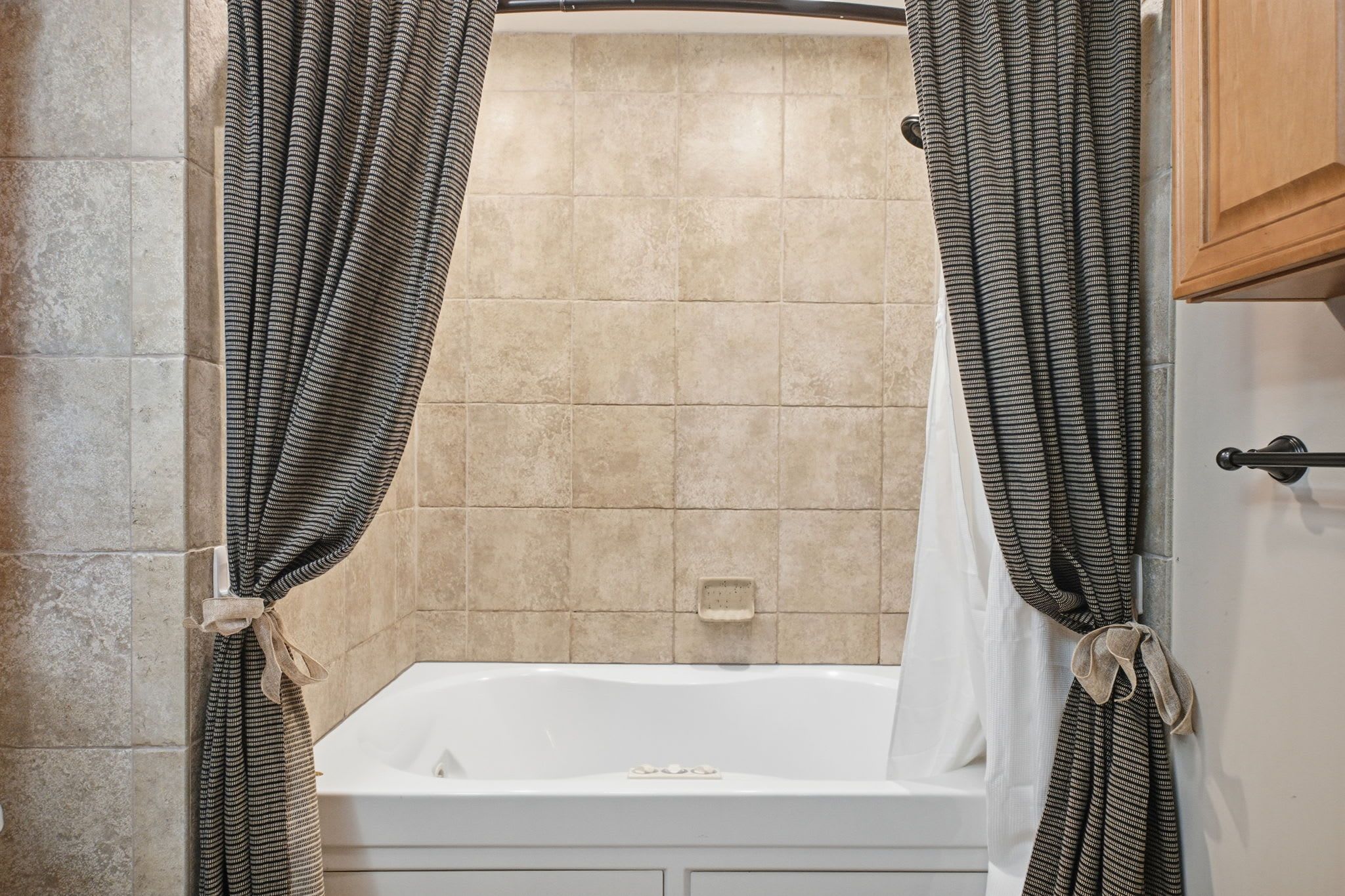 A bathroom with a bathtub, beige tiled walls, and patterned curtains tied back.