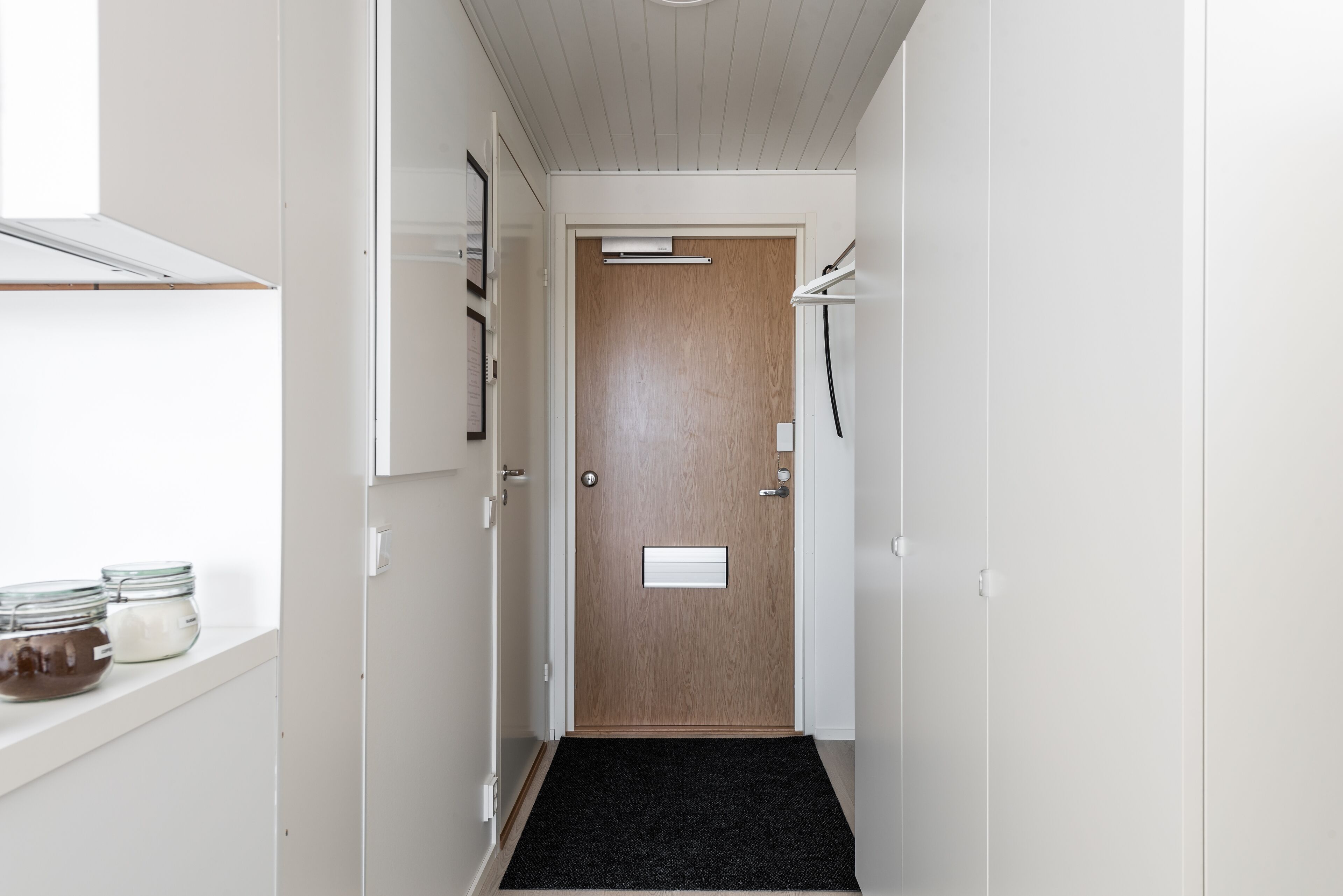 A narrow hallway with a wooden door, a black carpet, and a shelf with jars.