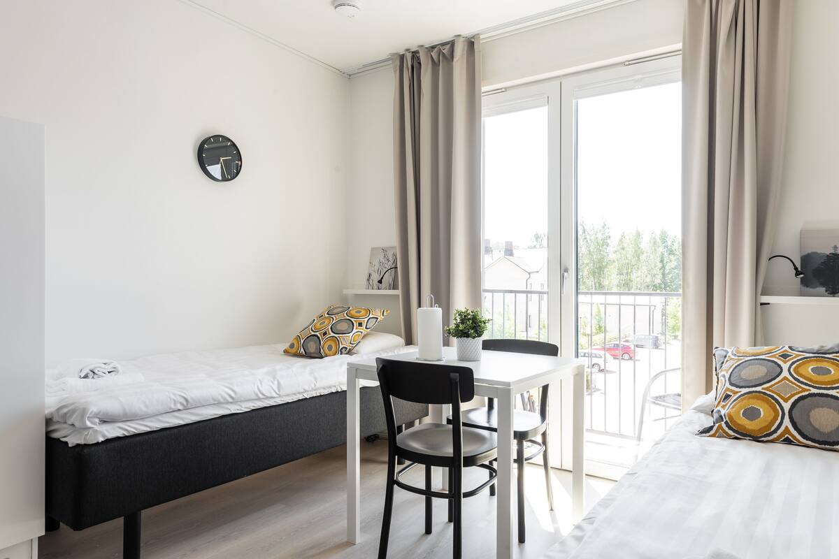A compact room with a bed, a small table with a chair, a clock on the wall, and a balcony with a view of the outside.