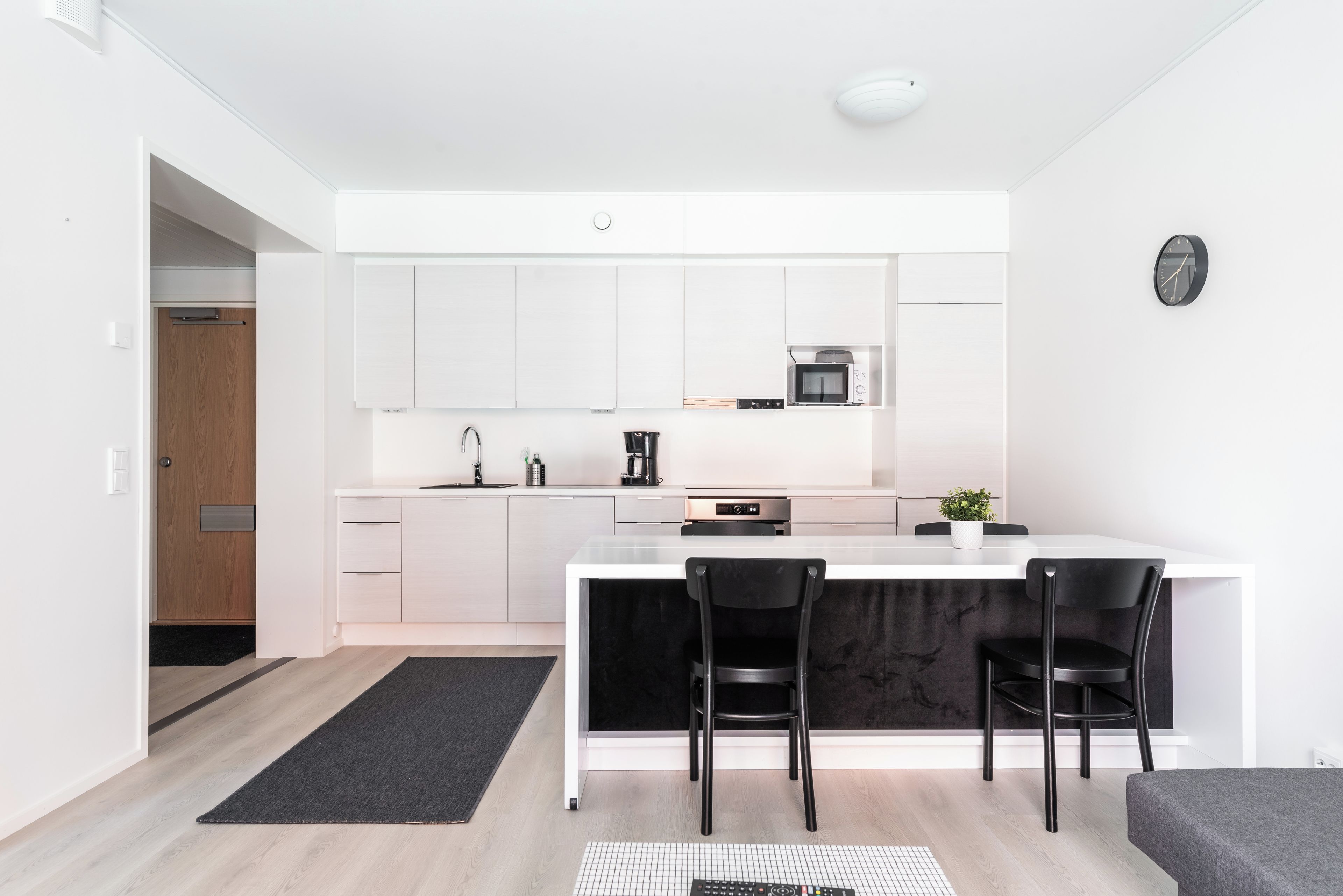 A modern kitchen with white cabinetry, a black countertop, and a built-in microwave. There are two black chairs and a small potted plant on the counter.