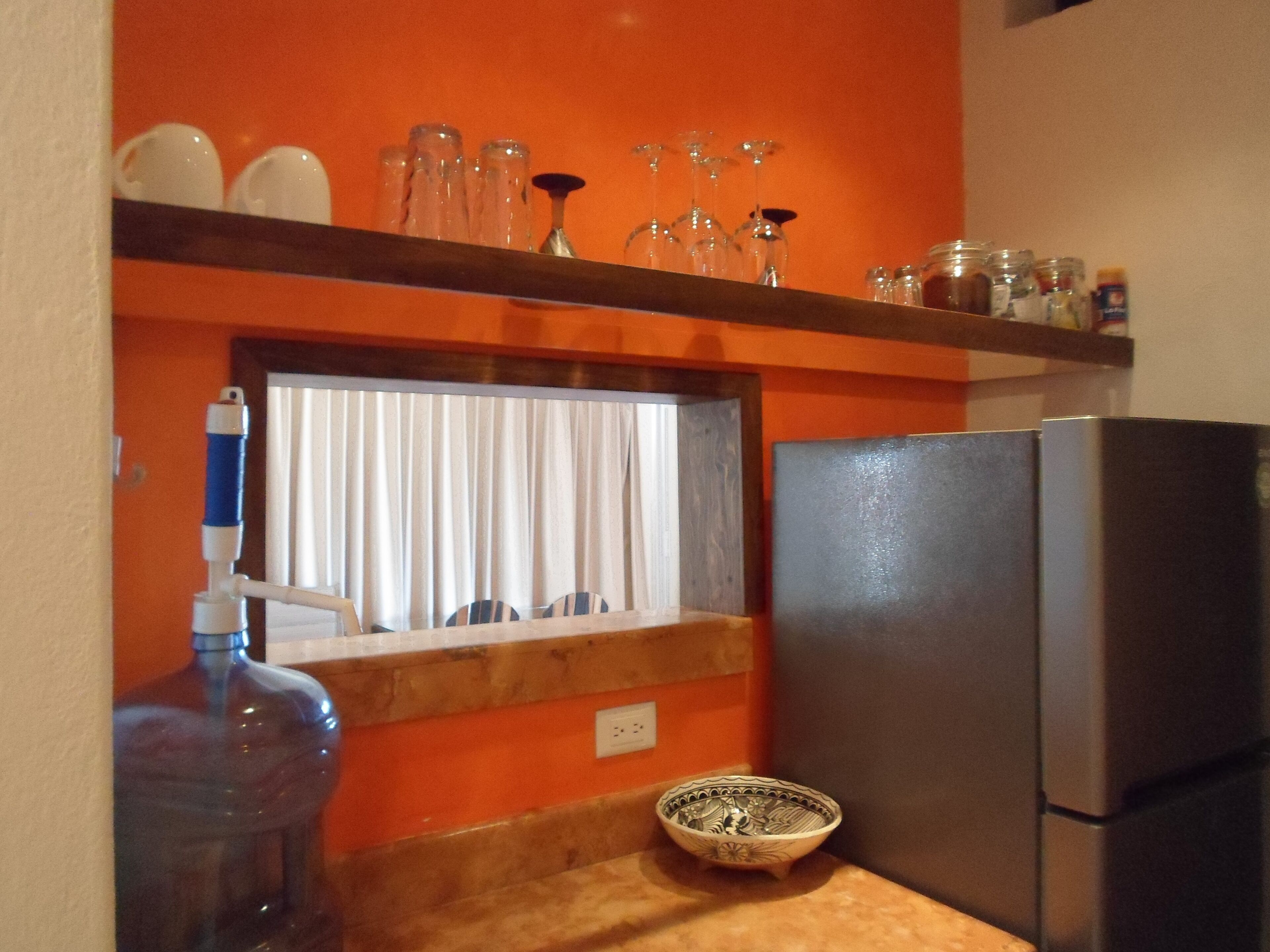A kitchen with a refrigerator, a water dispenser, a window with blinds, and a shelf with glassware and jars.
