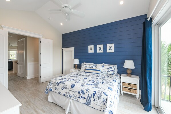 Un dormitorio con pared azul, una cama con cubrecama de motivos marinos en azul y blanco, dos mesitas de noche con lámparas y un ventilador de techo.