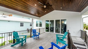 A covered porch with wooden flooring and a ceiling fan, featuring blue Adirondack chairs and a staircase leading to a glass door entrance.