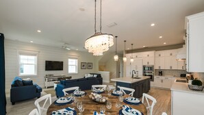 A modern kitchen with white cabinetry, a dining area with a wooden table set for six, and a living area with a dark blue sofa and a flat-screen TV.