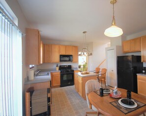 A compact kitchen with wooden cabinets, a black refrigerator, a microwave, and a stove. There is a dining table with chairs and a hanging light fixture.