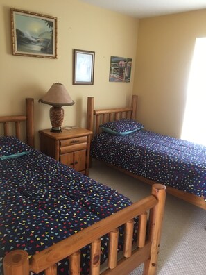 A room with two wooden beds, a wooden nightstand with a lamp, and a small drawer. There are framed pictures on the wall.