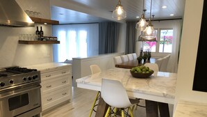 Een moderne keuken met een centraal kookeiland, roestvrijstalen apparatuur en een eethoek met witte stoelen en een schaal fruit.
