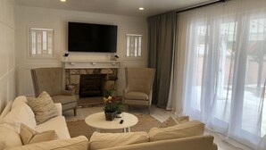 Een woonkamer met een open haard, twee fauteuils, een bank, een flatscreen-televisie en een groot raam met gordijnen.