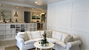 Een moderne woonkamer met een witte hoekbank, een kleine witte salontafel, en een keuken op de achtergrond.
