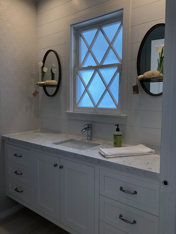 A bathroom with a white vanity, a window with a diamond pattern, a round mirror, and a towel on the counter.