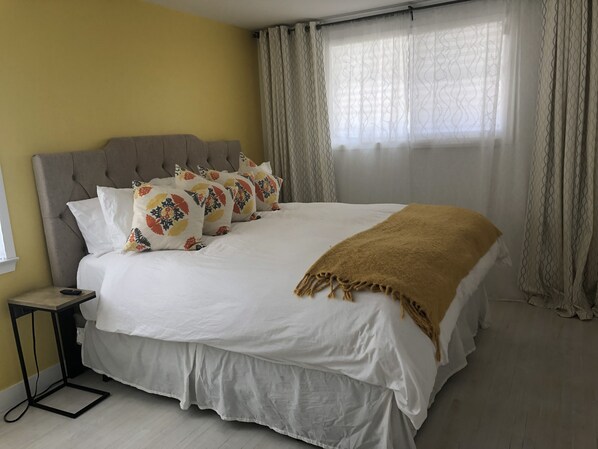 Una cama cuidadosamente tendida con edredón blanco y una manta marrón, un cabecero tapizado en gris y almohadas estampadas.