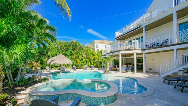 Una piscina con sillones de descanso y una sombrilla de paja, rodeada de palmeras y un edificio de varios pisos con balcones.