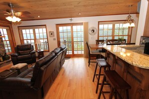 A spacious living area with a fireplace, leather seating, wooden flooring, and large windows offering a view of the outdoors.