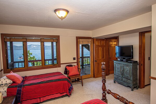 A bedroom with a bed, a television, a wooden cabinet, and a view of the water through large windows.