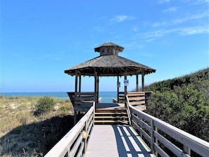 A wooden boardwalk leading to a gazebo with a clock on top, overlooking a beach.