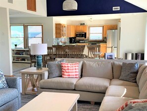 A living room with a sofa, a coffee table, and a lamp. There is a kitchen in the background with cabinets and appliances.