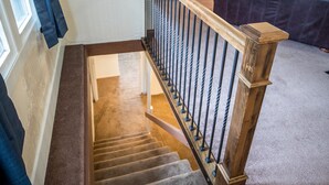 Una escalera empapelada con barandilla de madera y balaustres negros, que conduce a un salón con un sofá de color oscuro y un televisor.