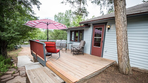 Una terraza de madera con muebles de exterior, un paraguas a rayas rojo y blanco, y una casa con puerta y ventanas rojas.