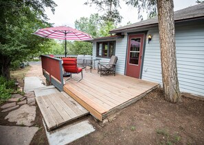 Une terrasse en bois avec des meubles d’extérieur, un parasol à rayures rouges et blanches, et une maison avec une porte et des fenêtres rouges.