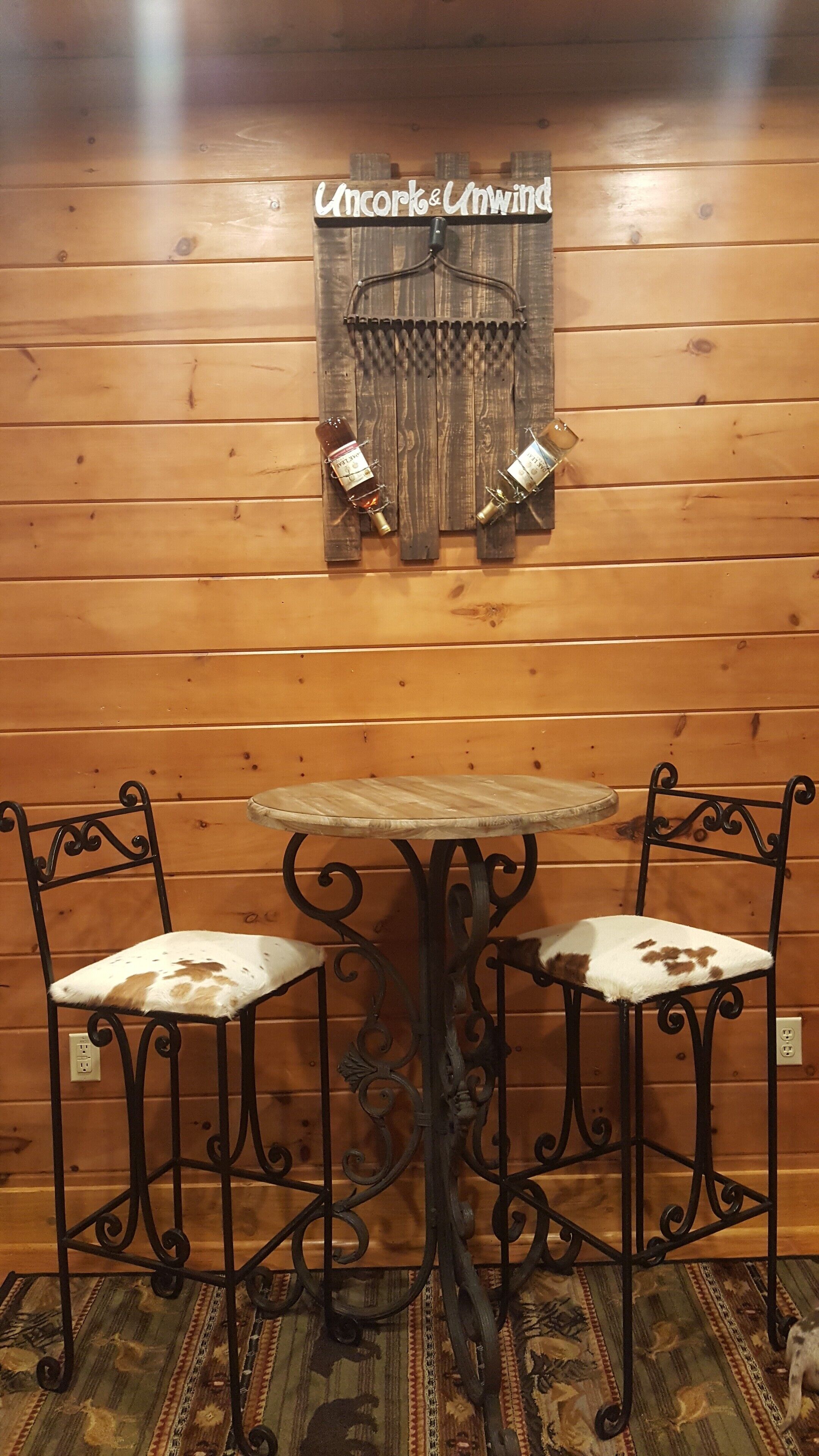 A wooden interior with a wooden wall, a sign reading "Uncork & Unwind", two metal chairs with white cushioned seats, and a small table with a metal base.