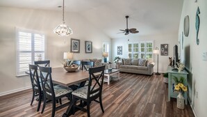 Uma sala de estar espaçosa com uma mesa de jantar de madeira, cadeiras escuras, um sofá e um ventilador de teto.