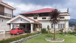 A two-story house with a red roof, a covered driveway, and a well-maintained garden with a palm tree and a circular stone border. - GuestHouser 3 BHK Cottage 563f (Ootacamund)