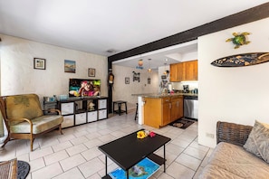 A living room with a sofa, a coffee table, and a television. There is a kitchen area with wooden cabinets and a countertop.
