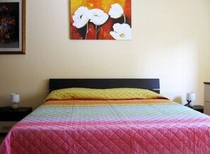 Una cama con un edredón de colores de arcoíris y una almohada amarilla, un cuadro de flores blancas en la pared, y una mesita de noche con una lámpara.