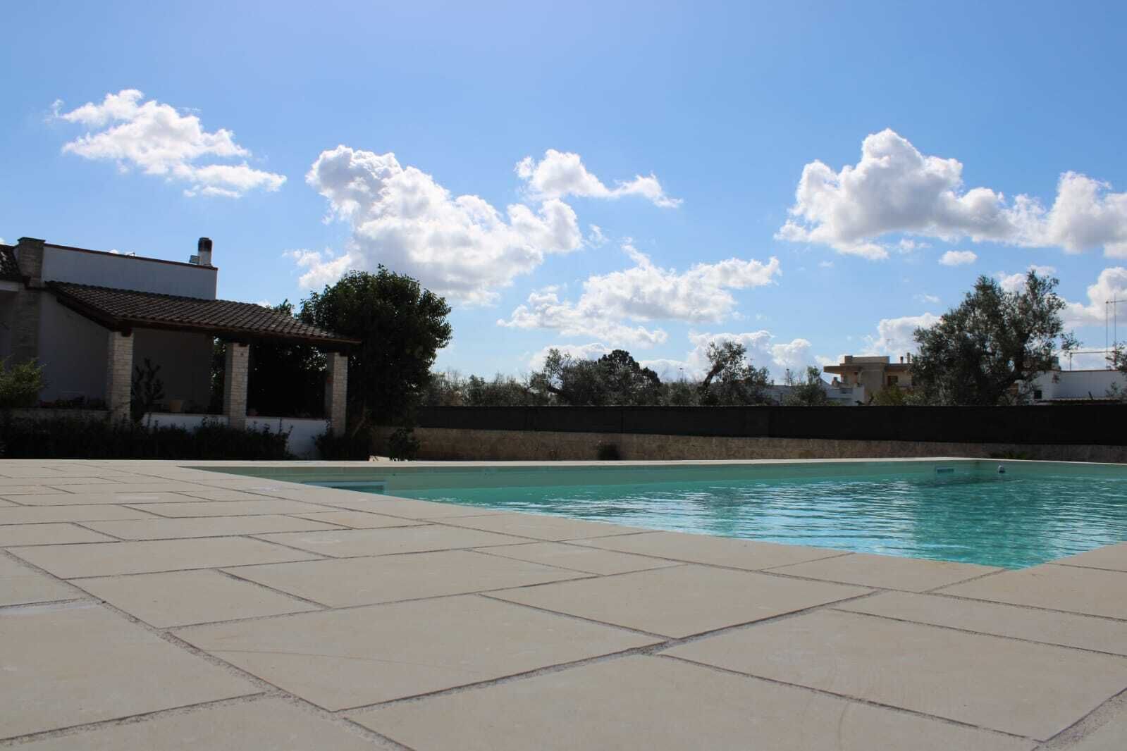 Une piscine dotée d’une terrasse carrelée, entourée d’un petit muret en pierre, sous un ciel dégagé parsemé de quelques nuages.