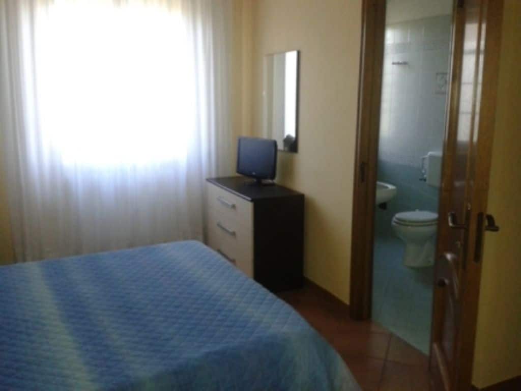 Een slaapkamer met een bed, een bureau met een computer, een spiegel en een badkamer met toilet en wastafel.