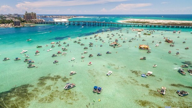 Eine Luftaufnahme einer Küstenlandschaft mit zahlreichen Booten in klarem türkisblauem Wasser, einer in die See führenden Mole und einem weit entfernten Gebäudekomplex.