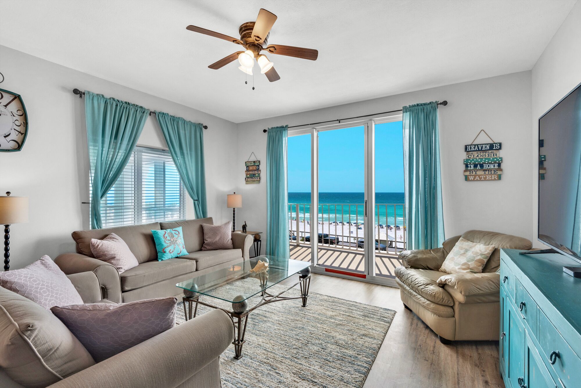 A living room with a view of the beach, a ceiling fan, and a clock on the wall.